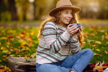 Sıcak giysili genç bir kadın piknikte ormanda oturur ve termostan sıcak çay doldurur. Doğa, yürüyüş, aktif yaşam tarzı, kamp..