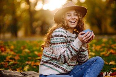 Sıcak giysili genç bir kadın piknikte ormanda oturur ve termostan sıcak çay doldurur. Doğa, yürüyüş, aktif yaşam tarzı, kamp..