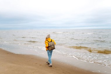 Bulutlu bir günde, turist bir kadının kumsalda yürüyüşünün arka görüntüsü. Soğuk hava. Sakinlik ve sükunet. Seyahat, turizm konsepti.