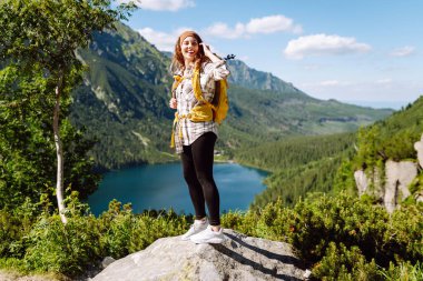 Genç kadın yürüyüşçü kayanın üzerinde duruyor ve dağların manzarasından hoşlanıyor. Aktif yaşam tarzı. Seyahat, macera.
