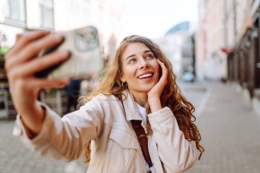 Şehir sokaklarında selfie çekerken kameraya gülümseyen iyimser genç bir kadın. Teknoloji, blog yazma, iletişim kavramı.