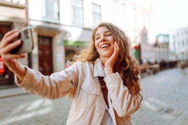 Şehir sokaklarında selfie çekerken kameraya gülümseyen iyimser genç bir kadın. Teknoloji, blog yazma, iletişim kavramı.