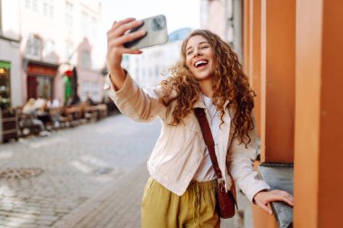 Şehir sokaklarında selfie çekerken kameraya gülümseyen iyimser genç bir kadın. Teknoloji, blog yazma, iletişim kavramı.
