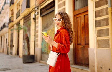 Genç bir kadın şehir merkezinde telefon yürüyüşü yapıyor. Teknoloji, bağlantı, blog yazma, modern yaşam tarzı kavramı.