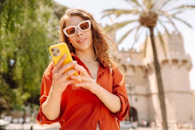 Genç bir kadın şehir merkezinde telefon yürüyüşü yapıyor. Teknoloji, bağlantı, blog yazma, modern yaşam tarzı kavramı.