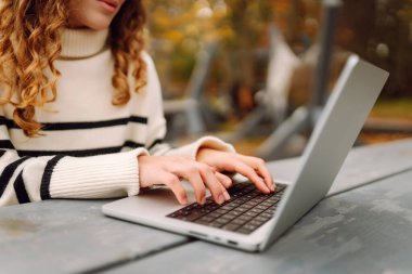 Kıvırcık saçlı genç bir kadın sonbahar parkında dizüstü bilgisayar kullanıyor. Açık havada çalışan serbest çalışan genç bir kadının portresi. E-öğrenme konsepti. Uzak çalışma.