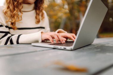 Kıvırcık saçlı genç bir kadın sonbahar parkında dizüstü bilgisayar kullanıyor. Açık havada çalışan serbest çalışan genç bir kadının portresi. E-öğrenme konsepti. Uzak çalışma.