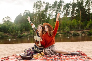 Kaygısız kadınlar plajda piknik yapıyor. Sonbahar havası. Pozitif modeller ekose üzerinde oturuyor, kahve içiyor, meyve yiyor. İnsanlar, yaşam tarzı, rahatlama ve tatil kavramı.