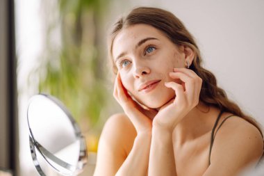 Kadın, sivilce sorununun mini aynayla oluştuğunu görünce yüzü için endişeleniyor. Sivilceler, sivilceler, gözenekler, yaralar. Filtresiz doğal cilt