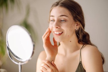 Kadın, sivilce sorununun mini aynayla oluştuğunu görünce yüzü için endişeleniyor. Sivilceler, sivilceler, gözenekler, yaralar. Filtresiz doğal cilt