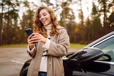 Arabanın yanında duran ve telefonu kullanan genç bir kadın. Araba paylaşımı, kiralama servisi ya da taksi uygulaması. Yaşam tarzı, seyahat, turizm, aktif yaşam. 
