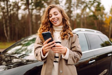 Arabanın yanında duran ve telefonu kullanan genç bir kadın. Araba paylaşımı, kiralama servisi ya da taksi uygulaması. Yaşam tarzı, seyahat, turizm, aktif yaşam. 