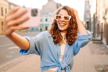 Selfie zamanı. Saçları bukleli, sokakta poz veren tasasız bir kadın. Açık havada pozitif model. Kentsel yaşam tarzı konsepti
