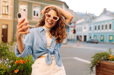 Selfie zamanı. Saçları bukleli, sokakta poz veren tasasız bir kadın. Açık havada pozitif model. Kentsel yaşam tarzı konsepti