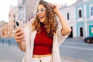 Genç bir kadın sokakta yürüyor ve akıllı telefon kamerasıyla selfie çekiyor. Açık havada pozitif model. Seyahat, blog, hafta sonu, rahatlama ve yaşam tarzı