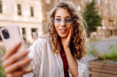 Genç bir kadın sokakta yürüyor ve akıllı telefon kamerasıyla selfie çekiyor. Açık havada pozitif model. Seyahat, blog, hafta sonu, rahatlama ve yaşam tarzı