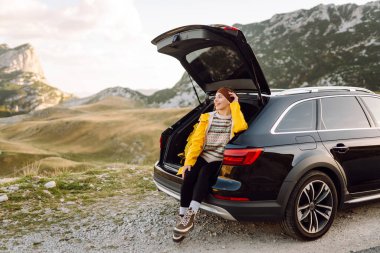 Genç bir kadın bir arabanın bagajında oturuyor. Yolculuk sırasında dur. Arabayla seyahat, turizm, gezi, aktif yaşam tarzı..