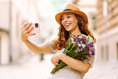 Selfie zamanı. Elinde bir buket kır çiçeğiyle sokakta poz veren genç bir kadın. Blog konsepti. Moda tarzı.