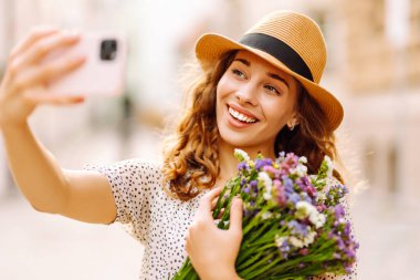 Selfie zamanı. Elinde bir buket kır çiçeğiyle sokakta poz veren genç bir kadın. Blog konsepti. Moda tarzı.