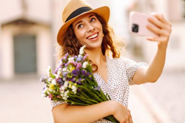 Selfie zamanı. Elinde bir buket kır çiçeğiyle sokakta poz veren genç bir kadın. Blog konsepti. Moda tarzı.