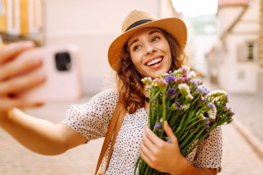 Selfie zamanı. Elinde bir buket kır çiçeğiyle sokakta poz veren genç bir kadın. Blog konsepti. Moda tarzı.