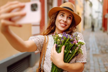 Selfie zamanı. Elinde bir buket kır çiçeğiyle sokakta poz veren genç bir kadın. Blog konsepti. Moda tarzı.