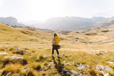 Genç bir kadın kuru otların arasında bir patika boyunca yürüyor. Güneşli bir günde dağlarla kaplı güzel bir manzara. Yürüyüş. Etkin yaşam biçimi