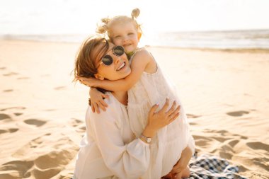 Neşeli bir anne sıcak bir yaz öğleden sonrasında güneşli bir plajda kızını kucaklıyor, aşk ve neşe dolu bir anı yakalıyor.