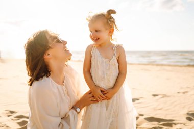 Neşeli bir anne sıcak bir yaz öğleden sonrasında güneşli bir plajda kızını kucaklıyor, aşk ve neşe dolu bir anı yakalıyor.