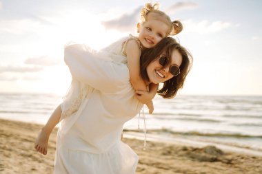 Neşeli bir anne sıcak bir yaz öğleden sonrasında güneşli bir plajda kızını kucaklıyor, aşk ve neşe dolu bir anı yakalıyor.