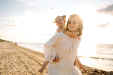 Neşeli bir anne sıcak bir yaz öğleden sonrasında güneşli bir plajda kızını kucaklıyor, aşk ve neşe dolu bir anı yakalıyor.