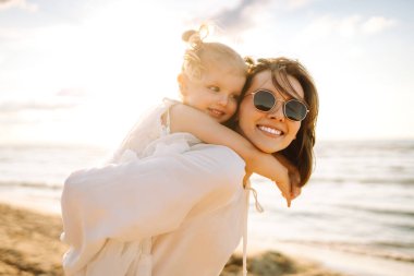 Neşeli bir anne sıcak bir yaz öğleden sonrasında güneşli bir plajda kızını kucaklıyor, aşk ve neşe dolu bir anı yakalıyor.