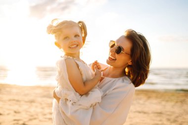 Neşeli bir anne sıcak bir yaz öğleden sonrasında güneşli bir plajda kızını kucaklıyor, aşk ve neşe dolu bir anı yakalıyor.