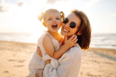 Neşeli bir anne sıcak bir yaz öğleden sonrasında güneşli bir plajda kızını kucaklıyor, aşk ve neşe dolu bir anı yakalıyor.