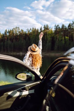 Neşeli bir gezgin gün batımında göl kenarında açık bir araba kapısı ile doğayı kucaklıyor, özgürlük ve macera yayıyor.
