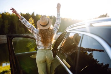 Neşeli bir gezgin gün batımında göl kenarında açık bir araba kapısı ile doğayı kucaklıyor, özgürlük ve macera yayıyor.