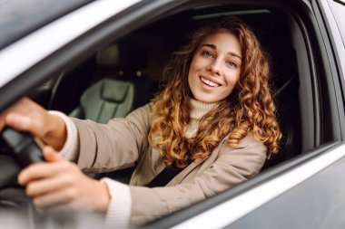 Açık güneşli bir günde, şehirde araba kullanırken gülümseyen kıvırcık saçlı genç bir kadın.