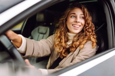 Açık güneşli bir günde, şehirde araba kullanırken gülümseyen kıvırcık saçlı genç bir kadın.