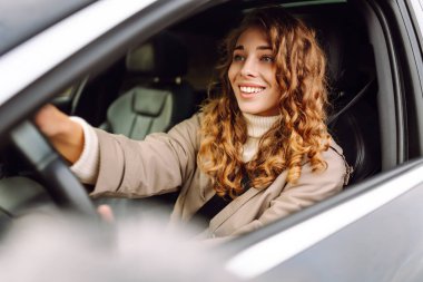 Açık güneşli bir günde, şehirde araba kullanırken gülümseyen kıvırcık saçlı genç bir kadın.