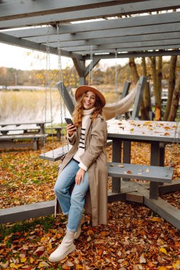 Açık havada huzurlu bir sonbahar gününün tadını çıkaran genç bir kadın telefonu renkli yapraklarla çevrili bir masada oturuyor.