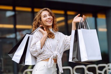Alışveriş torbalarıyla neşeli genç bir kadın canlı bir şehir ortamında modern bir tuğla binanın dışında güneşli bir günün tadını çıkarıyor.