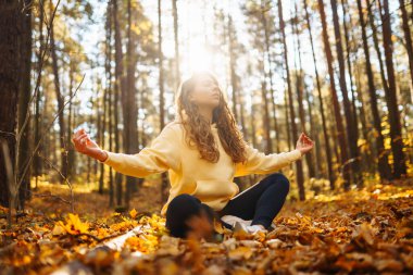 Genç bir kadın, öğleden sonra saatlerinde sakin bir ormanda altın sonbahar yaprakları arasında farkındalık meditasyonu yapar.