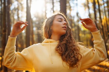 Genç bir kadın, öğleden sonra saatlerinde sakin bir ormanda altın sonbahar yaprakları arasında farkındalık meditasyonu yapar.