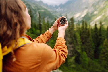 Sırt çantalı genç bir kadın, açık mavi gökyüzünün altında yüksek ağaçlar ve geniş dağlarla çevrili kayalık bir çıkıntının üzerinde dururken pusulasını inceliyor..