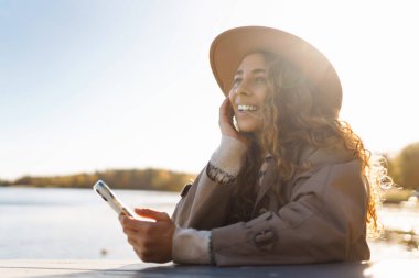 Sonbaharda güneşli bir parkta akıllı telefon kullanarak dışarıda kahve içen genç bir kadın.