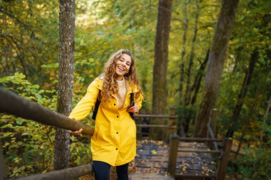 Sarı yağmurluklu bir kadın, rengarenk bir sonbahar ormanında ahşap bir yolda yürürken doğaya hayran kalır.