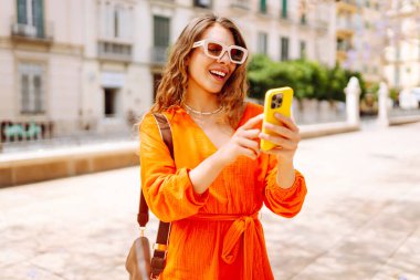 Güneşli bir günün tadını çıkaran bir kadın tarafından giyilen parlak turuncu elbise. Resimli bir şehir ortamında selfie çekerken.