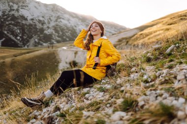 Sarı ceketli neşeli bir kadın güneşli bir günde arka planda dağlarla bir yamaçta duruyor.