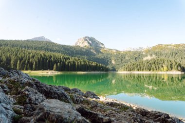 Berrak turkuaz göl yemyeşil ormanlarla ve güneşli bir günde parlak bir gökyüzünün altında dağlarla çevrili.