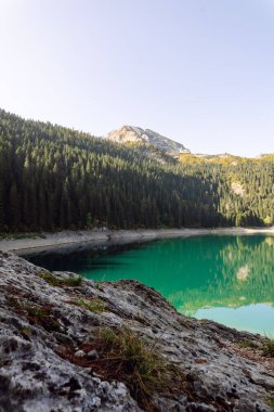 Berrak turkuaz göl yemyeşil ormanlarla ve güneşli bir günde parlak bir gökyüzünün altında dağlarla çevrili.